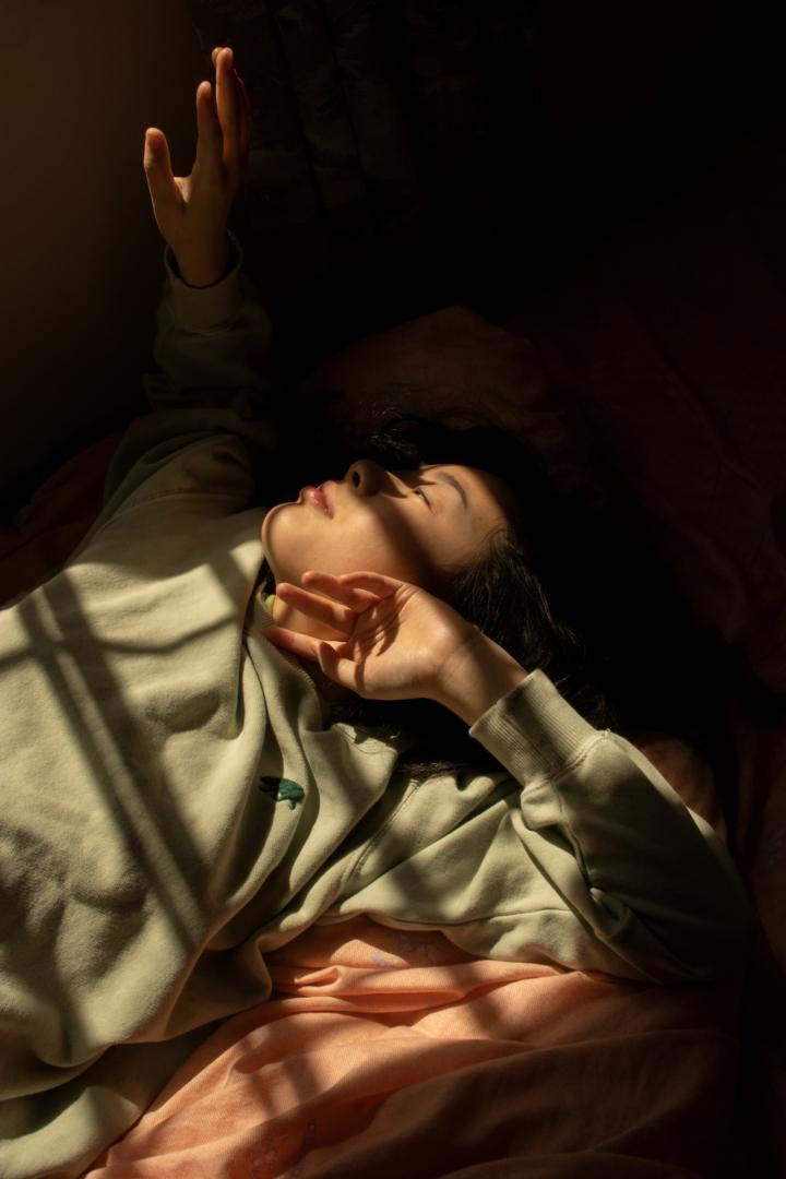 A woman lays in bed while the shadows of a window stretch over her face. 